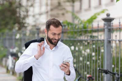 Smiling Handsome Man with Coat on the Shoulder Using Mobile Phone While Walking on the Street